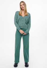 Groene gebreide cardigan met een V-hals en vijf knopen, gecombineerd met groene wijde broek en zwarte schoeisel. Eenvoudig, textuurontwerp.
