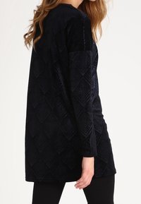 Czarna, welwetowa tunika z długim rękawem, ozdobiona teksturowym wzorem w romby i luźnym krojem. Gładki materiał z delikatnym połyskiem i bocznymi rozcięciami.