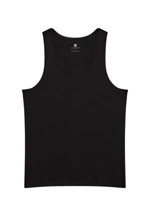 Ung mand står barfodet med hænderne i lommerne iført en sort tanktop og sorte joggebukser mod en ensfarvet lys baggrund.