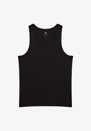 Ung mand står barfodet med hænderne i lommerne iført en sort tanktop og sorte joggebukser mod en ensfarvet lys baggrund.