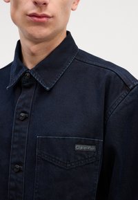 Camisa de denim escuro com fecho de botão, colarinho pontudo, um bolso no peito e uma etiqueta com o logo Calvin Klein no bolso. Tecido texturizado.
