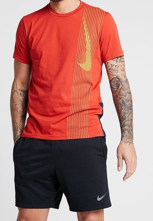 Mężczyzna noszący czerwoną koszulkę Nike z żółtym logo i czarne spodenki Nike, stojący z rękami w kieszeniach, pokazujący tatuaże na obu przedramionach.