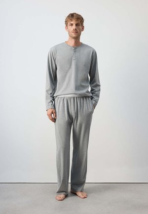 Mann steht barfuß, trägt ein hellgraues langärmeliges Henley-Shirt und passende locker sitzende Hosen vor einem einfarbigen Hintergrund.