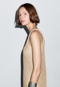 Beige strikket tanktop med løst, tekstureret design, der viser brede skulderstropper. Sølvfarvet metallisk armbånd på håndleddet.