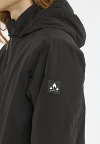 Schwarze wasserdichte Jacke mit Kapuze, featuring eine Stoffstruktur und ein Logo-Patch auf dem Ärmel. Saubere Linien und minimalistisches Design.
