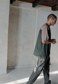 Mann in lockerem zweifarbigen Hemd und grauer Hose geht drinnen an einer schlichten weißen Wand mit hölzernen Deckenbalken darüber entlang.