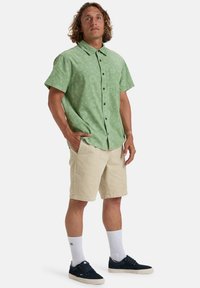 Groene kortlopende button-up shirt met een geometrisch patroon, gecombineerd met beige shorts, witte sokken en marineblauwe sneakers.