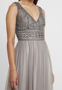 Beaded evening dress in light gray, featuring a V-neck and a fitted bodice. The skirt has flowing layers of sheer fabric with a textured waistband.