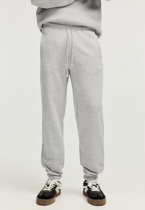 Person iført lysegrå sweatpants med snøre og sorte sneakers med hvide snørebånd, stående mod en ensfarvet baggrund.