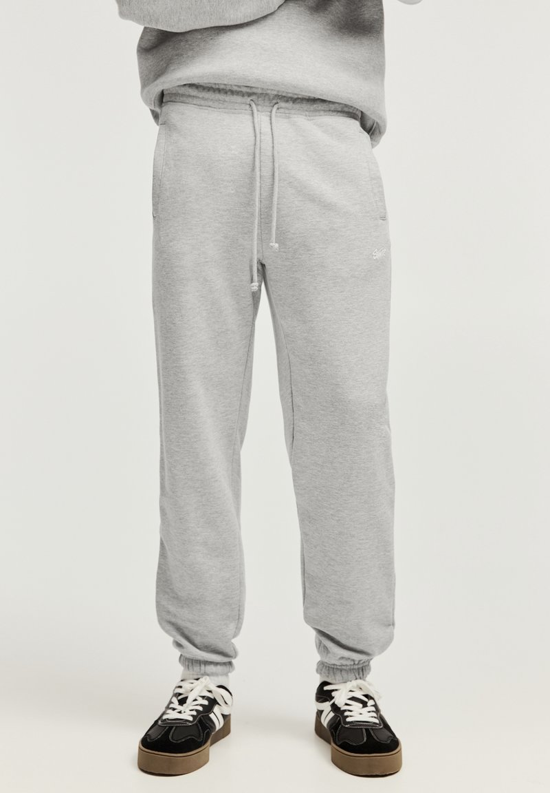 Personne portant un pantalon de survêtement gris clair avec un cordon de serrage et des baskets noires à lacets blancs, debout devant un fond uni.