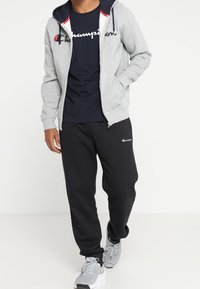 Grå dragkedja-hoodie med marinblå foder, vit logotyp; marinblå t-shirt, svarta sweatpants och grå sneakers med vita accenter. Avslappnad sportklädsel.