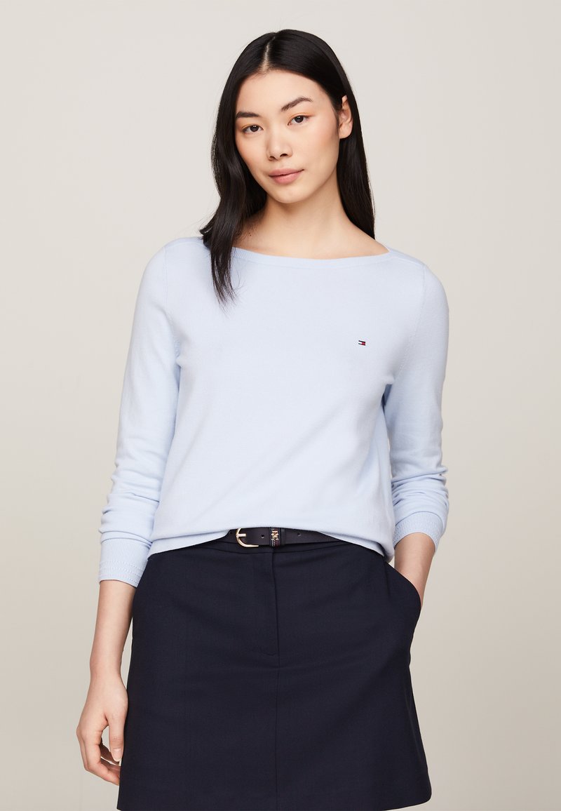 Maglione azzurro chiaro, con scollatura rotonda e maniche lunghe. Piccolo logo sul petto. Abbinato a una gonna scura e una cintura. Tessuto dalla consistenza liscia.