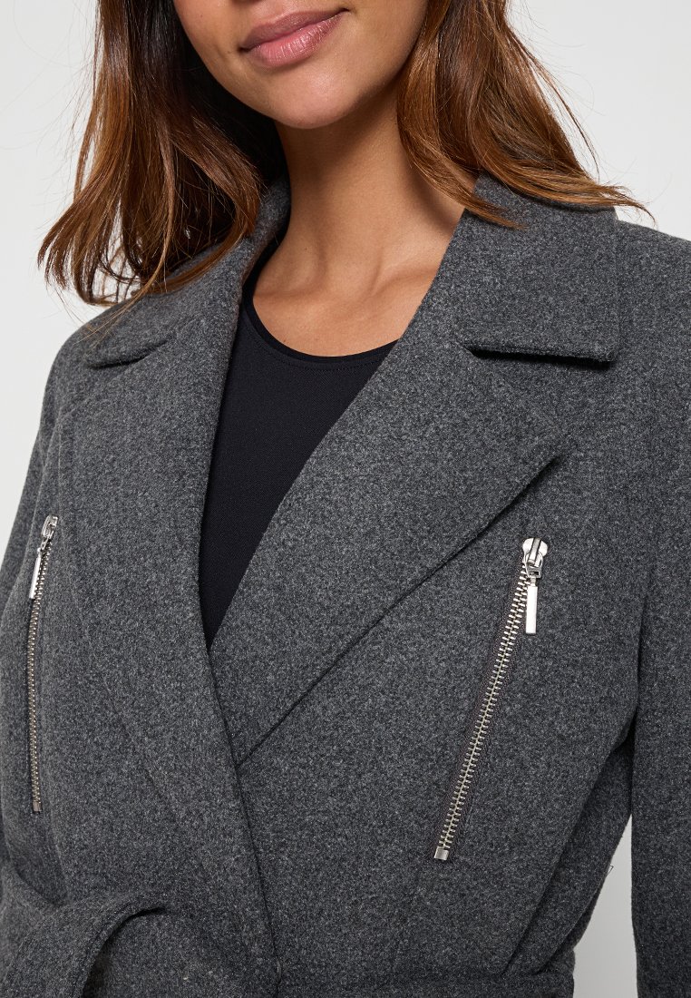 Femme portant un manteau en laine gris foncé avec des revers larges et des poches zippées verticales en argent sur un haut noir, montrée des épaules à la moitié du torse.