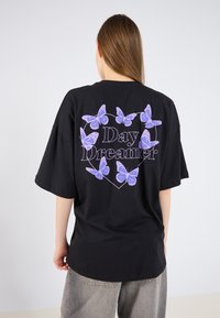 Sort oversized t-shirt med et hjerteformet design, der har lilla sommerfugle og teksten "Day Dreamer" i hvid. Korte ærmer, lavet af bomuldsmateriale.