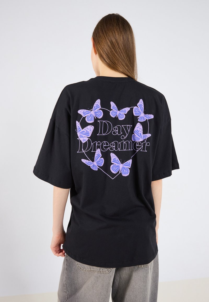 Sort oversized t-shirt med et hjerteformet design, der har lilla sommerfugle og teksten "Day Dreamer" i hvid. Korte ærmer, lavet af bomuldsmateriale.