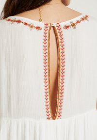 Valkoinen, ilmava mekko, jossa on syvä avoin selkä. Mekossa on oranssia kukkakirjontaa kaula-aukossa ja selässä. Napitusyksityiskohtia on näkyvissä.