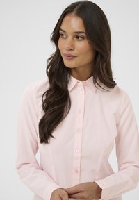 Chemise rose pâle à boutons, confectionnée dans un tissu doux, avec un col pointu, des manches longues et des accents de boutons discrets.