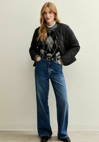Mujer con chaqueta acolchada negra, suéter gris de rombos, jeans de pierna ancha de tiro alto y zapatos negros de pie contra una pared sencilla.