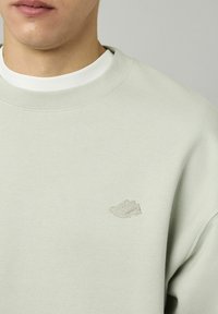 Sweatshirt vert clair avec un col rond, arborant un discret logo de basket brodé sur la poitrine. Texture de tissu douce et coupe décontractée.