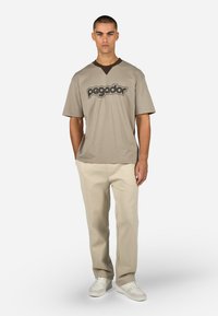 Beżowy t-shirt z krótkim rękawem z ciemnobrązowym dekoltem oraz logo z napisem "pegador". Połączony z jasnobeżowymi spodniami i białymi trampkami.