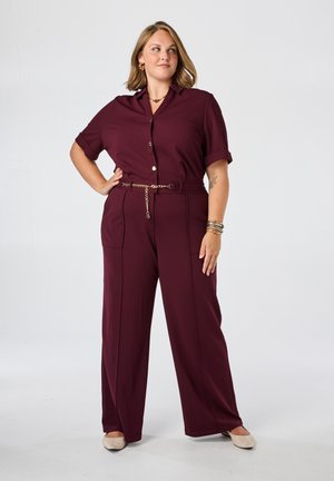 Vrouw staand met hand op heup, draagt een bordeauxrode korte mouw button-up jumpsuit en beige platte schoenen tegen een effen achtergrond.