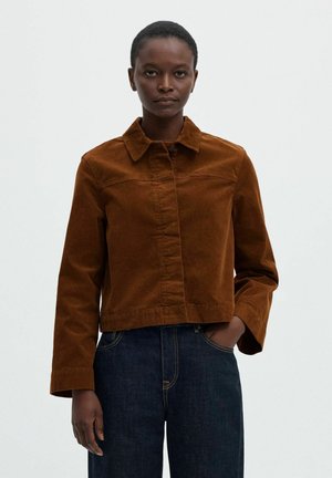 Femme portant une veste en velours côtelé marron avec col et un jean bleu foncé, debout avec une main dans la poche devant un fond uni.