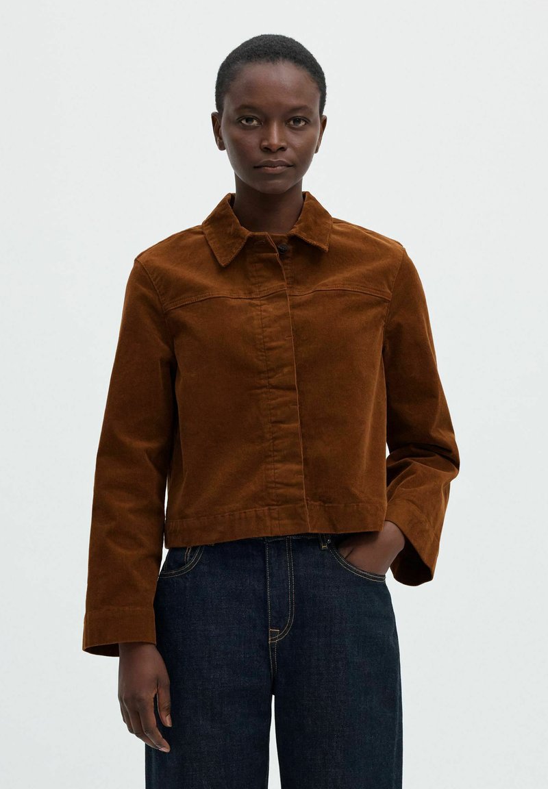 Femme portant une veste en velours côtelé marron avec col et un jean bleu foncé, debout avec une main dans la poche devant un fond uni.