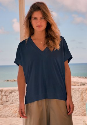 Joven mujer con cabello suelto y ondulado lleva blusa azul marino con cuello en V y pantalones beige, de pie al aire libre junto a una pared de piedra con el mar de fondo.