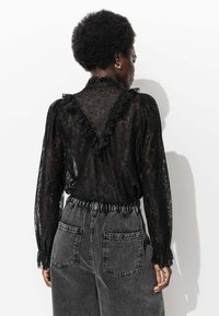 Blouse en dentelle noire avec un col et des poignets à volants, associée à un pantalon en denim gris taille haute avec une taille élastique et des poches arrière.