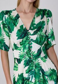 Robe florale verte et blanche avec des manches courtes bouffantes, un décolleté en V profond et des boutons décoratifs sur le devant. Texture de tissu lisse.