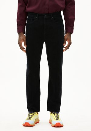 Pantalons en velours côtelé noir avec une coupe droite, dotés d'un style classique à cinq poches. Portés avec des chaussures de sport jaunes et grises.
