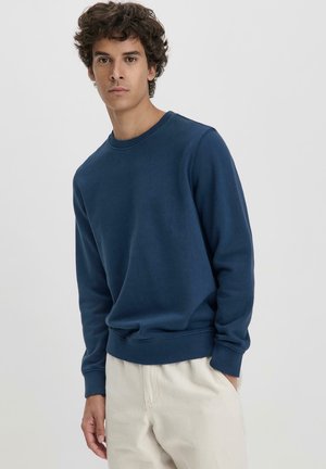 Navy blue sweatshirt in soft material, featuring a round neckline and ribbed cuffs and hem. Worn with light-colored trousers.