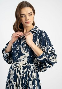Granatowa sukienka w kwiatowy wzór w kolorze kremowym, z kołnierzykiem, podkreśloną talią oraz bufkowymi rękawami, wykonana z lekkiego materiału.