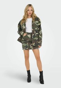 Sada kabátu a sukně s camo vzorem v zelené, hnědé a béžové barvě; odolný materiál; má kapsy; kombinováno s černými kotníkovými botami.