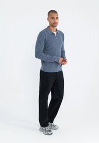 Sudadera azul claro con cremallera y textura acanalada, combinada con pantalones negros y zapatillas deportivas grises, llevada por un modelo que se encuentra de pie frente a un fondo blanco.
