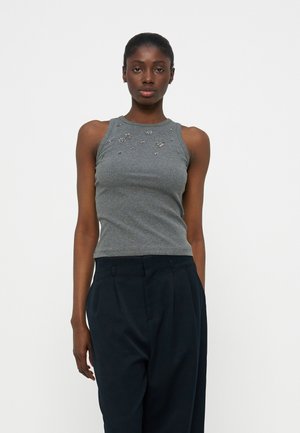 Γυναίκα που φοράει αμάνικη γκρι μπλούζα με μικρές ανθισμένες διακοσμήσεις και ψηλόμεσο μαύρο παντελόνι με πιέτες, στέκεται μπροστά σε λευκό φόντο.