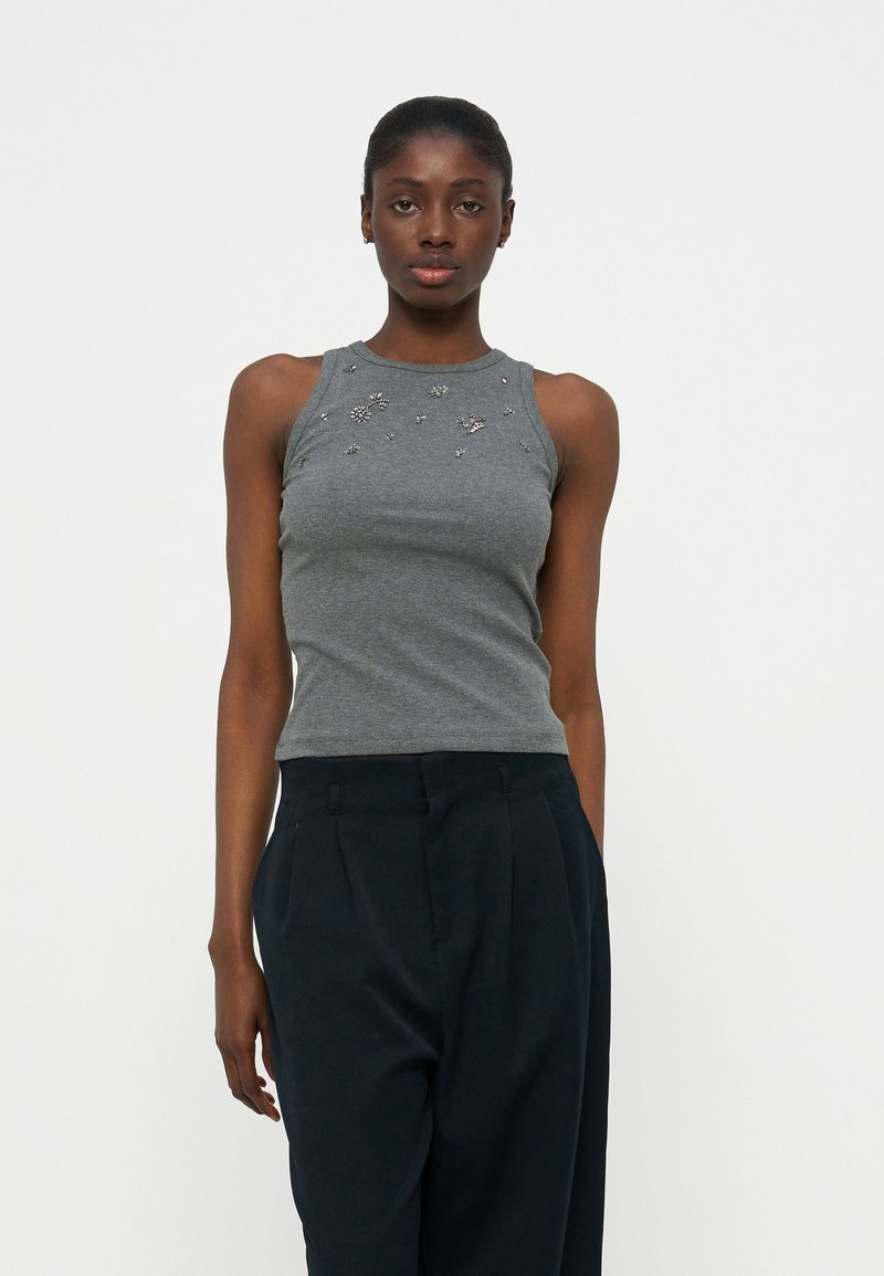 Γυναίκα που φοράει αμάνικη γκρι μπλούζα με μικρές ανθισμένες διακοσμήσεις και ψηλόμεσο μαύρο παντελόνι με πιέτες, στέκεται μπροστά σε λευκό φόντο.