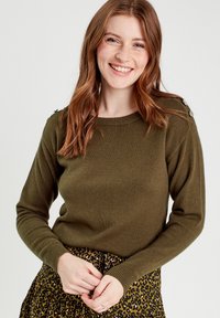 Olive green knit sweater with long sleeves and a round neckline, featuring decorative buttons on shoulders. Paired with a leopard print skirt.