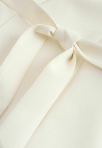 Cremefarbenes Stoffstück mit einem ordentlich gebundenen Knoten, der zwei breite Schlaufen und zwei herabhängende Enden bildet, wobei glatte Textur und Falten sichtbar sind.