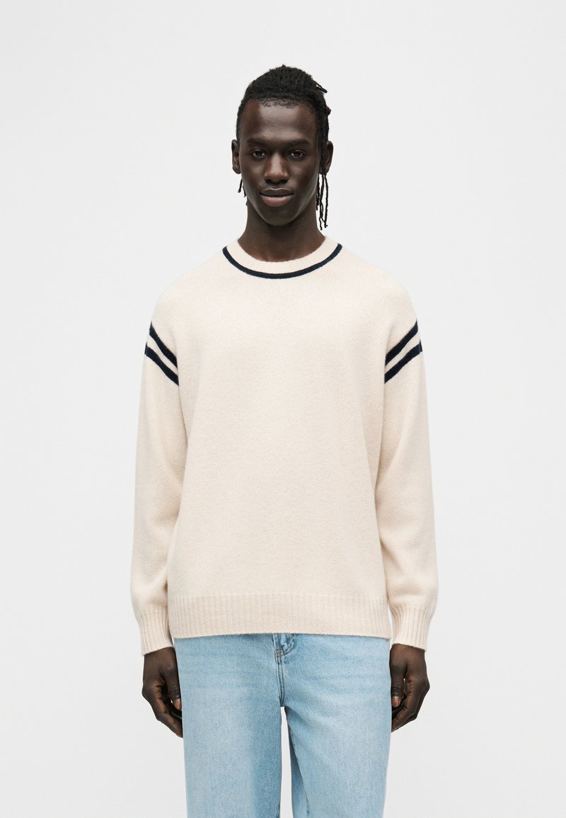 Πουλόβερ πλεκτό σε κρεμ χρώμα με μακριά μανίκια, μανσέτες ριμπ και τρεις μπλε σκούρες ρίγες στους ώμους. Το πουλόβερ έχει στρογγυλή λαιμόκοψη.