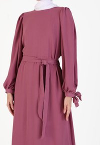 Robe à manches longues en rose poudre, avec des manches bouffantes, une taille nouée et des accents de nœuds aux poignets. Tissu doux et fluide avec une texture lisse.