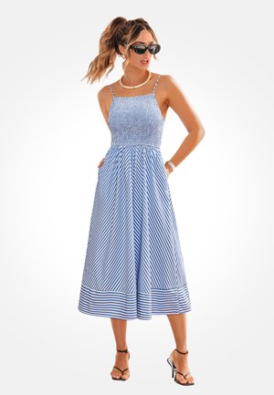 Femme avec queue de cheval portant des lunettes de soleil noires, une robe midi sans manches rayée bleu et blanc avec poches, un collier en or et des sandales à talons hauts noires.