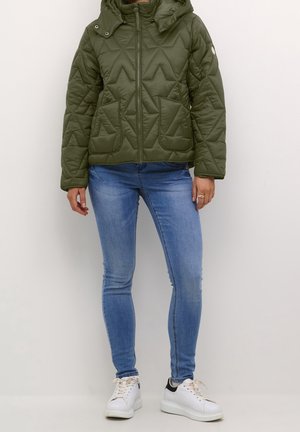 Osoba ubrana w oliwkowo-zieloną pikowaną kurtkę z kapturem, jasnoniebieskie obcisłe jeansy i białe trampki na jednolitym tle.