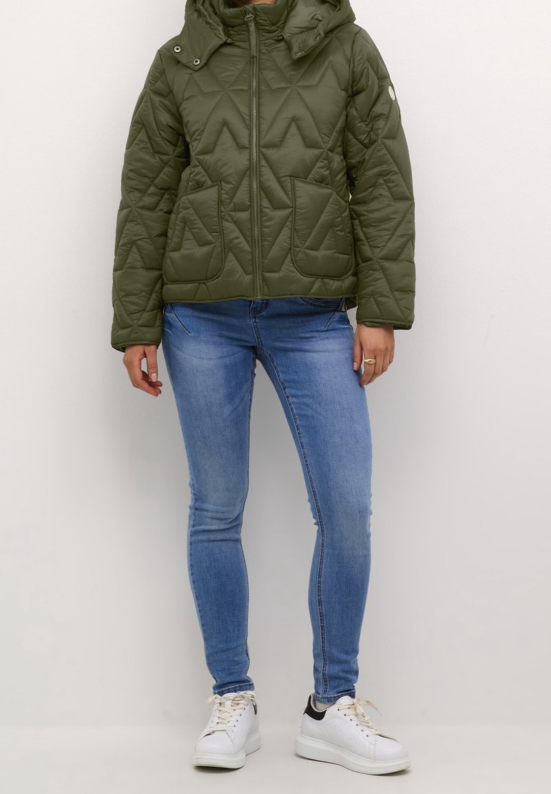 Personne portant une veste matelassée olive avec capuche, un jean skinny bleu clair et des baskets blanches sur un fond uni.