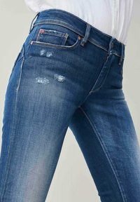 Jeans en denim bleu présentant une coupe slim, des zones délavées et des détails usés. Comprend une petite poche avant et des coutures contrastées.