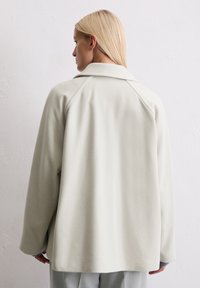 Veste en polaire gris clair avec un col à revers et des manches raglan. Le dos présente une surface lisse et texturée sans fermetures visibles.