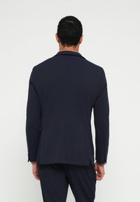 Σκούρο μπλε σακάκι από υφή υφάσματος· διαθέτει πέτο με χαραγή, δύο πλαϊνές σκισμές και κουμπιά στα μανίκια. Φαίνεται από πίσω.