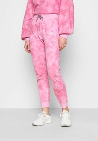 Kobieta ubrana w różowe spodnie dresowe w stylu tie-dye z elastycznymi mankietami i białymi trampkami, stojąca na jednolitym jasnym tle.