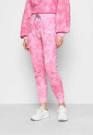 Nainen, joka on pukeutunut vaaleanpunaiseen tye-dye-housupukuun, jossa on joustavat hihnat, ja valkoisiin tennareihin, seisoo yksinkertaista vaaleaa taustaa vasten.