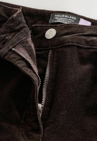 Dunkelbraune Cordhosen mit einem entspannten Bootcut-Design, ausgestattet mit einem silbernen Reißverschluss, einem Markenetikett und einem weichen, strukturierten Stoff.